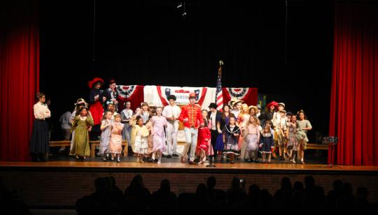 Music Man characters in 1912 clothing on stage dancing.