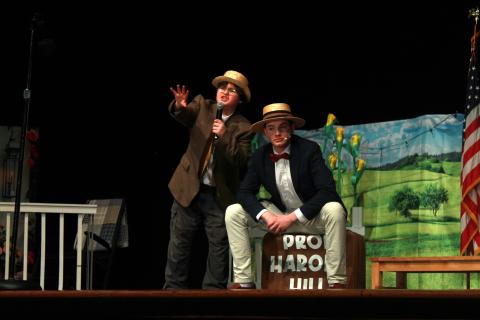 Two Music Man characters on stage, one sitting on a suitcase labeled 