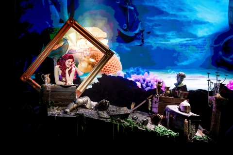 Photo Credit: Sydney Youth Musical Theatre A mermaid in a picture frame among a collection of undersea treasures against digital backdrop of the ocean floor