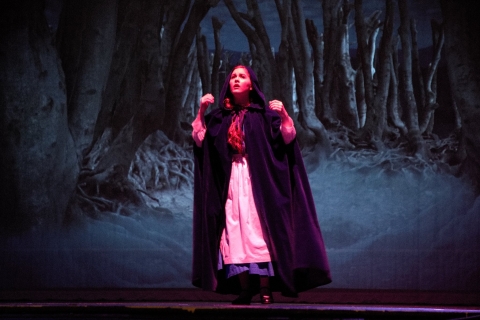 Photo Credit: Theatre Longview A girl wearing a cloak in front of a digital projection of a dark forest