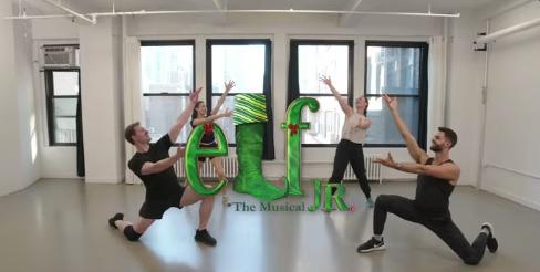 A group of three people with both their hands raised standing in a row, with a fourth person sitting on a chair on the far left.