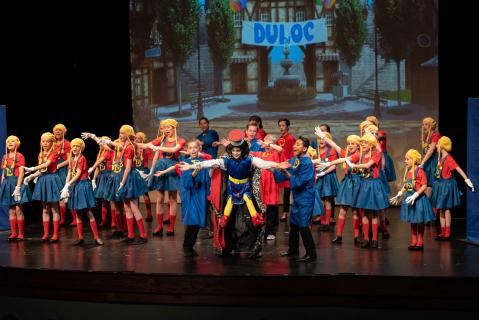 A group of performers in red tops and blue skirts with a performer in the middle wearing a blue velvet tunic in front of a digital projection of a town with a banner that reads 