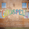 A curtain warmer of a brick building with windows and the Godspell logo in rainbow lettering
