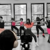 Five dancers wearing pink tops and shorts in a row demonstrating a dance move while someone is filming in front of them.