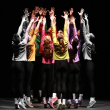 A group of dancers form a circle facing each other while reaching upward